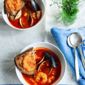 Two bowls of Julia Child's bouillabaisse in tomato broth, each topped with toasted bread and garnished with shellfish. Nearby, a small vase of fresh herbs and a blue napkin with silverware rest on a linen tablecloth.