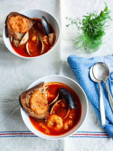 Two bowls of Julia Child's bouillabaisse in tomato broth, each topped with toasted bread and garnished with shellfish. Nearby, a small vase of fresh herbs and a blue napkin with silverware rest on a linen tablecloth.