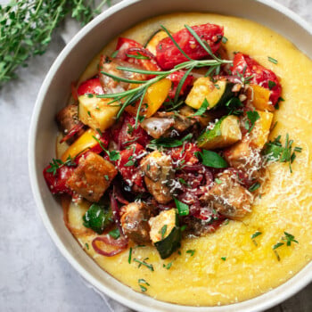 A bowl of creamy yellow polenta with roasted vegetables like zucchini, tomatoes, eggplant, and red peppers, finished with fresh herbs and grated cheese. A sprig of thyme is placed nearby for a touch of rustic charm.
