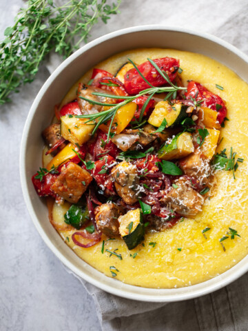 A bowl of creamy yellow polenta with roasted vegetables like zucchini, tomatoes, eggplant, and red peppers, finished with fresh herbs and grated cheese. A sprig of thyme is placed nearby for a touch of rustic charm.