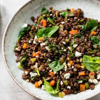 A bowl of lentil salad featuring spinach leaves, chopped carrots, crumbled feta cheese, and fresh herbs sits on a light textured surface.