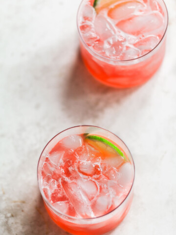 Two glasses filled with a pink rhubarb cocktail, each garnished with a lime wedge, are placed on a light-colored, textured surface.