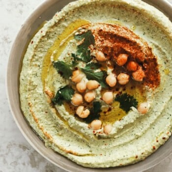A bowl of vibrant green chickpea hummus topped with whole chickpeas, paprika, olive oil, and fresh herbs sits on a light textured surface.