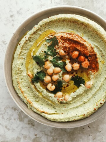 A bowl of vibrant green chickpea hummus topped with whole chickpeas, paprika, olive oil, and fresh herbs sits on a light textured surface.