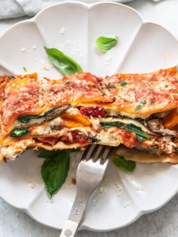 A slice of lasagna, showing layers of cheese, vegetables and tomato sauce, on a plate with a fork and fresh basil leaves.