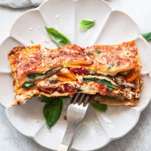 A slice of lasagna, showing layers of cheese, vegetables and tomato sauce, on a plate with a fork and fresh basil leaves.