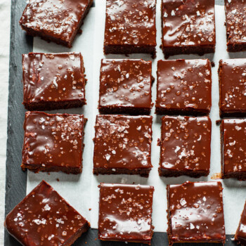 Sliced brownie squares on a slate board, topped with chocolate ganache frosting and flaked sea salt.