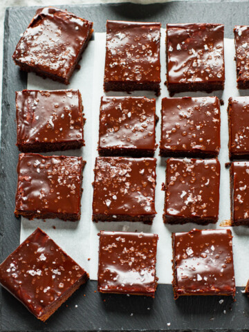 Sliced brownie squares on a slate board, topped with chocolate ganache frosting and flaked sea salt.
