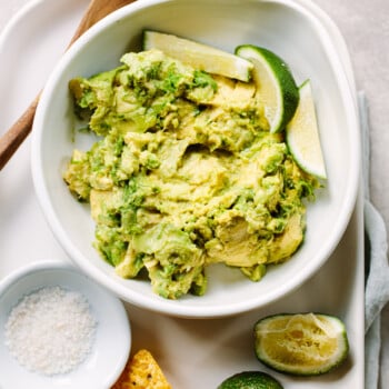 A bowl of avocado guacamole in a bowl, with tortilla chips, lime wedges and salt next to it.