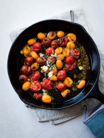 A black cast-iron skillet filled with roasted red and yellow cherry tomatoes, herbs, and sliced garlic sits on a folded gray kitchen towel—perfect for tossing into a vibrant cherry tomato pasta. The background is a light, textured surface.