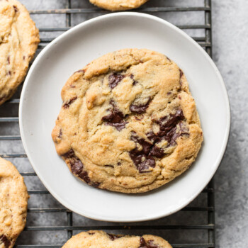 A chocolate chip cookie on a white plate sits on a cooling rack, surrounded by more chocolate chip cookies. The cookie is golden brown with visible chunks of melted chocolate.