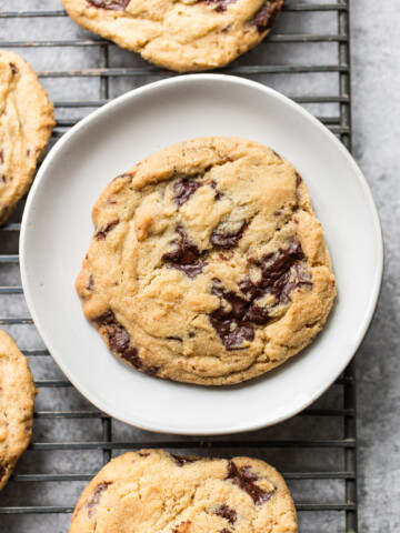 A chocolate chip cookie on a white plate sits on a cooling rack, surrounded by more chocolate chip cookies. The cookie is golden brown with visible chunks of melted chocolate.
