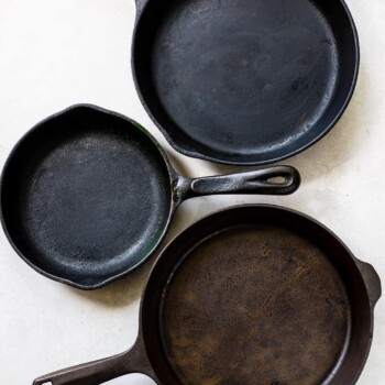 Three cast iron skillets of varying sizes are arranged on a white surface. Two have handles on one side, while the largest has two handles. Each skillet shows signs of use, revealing how to properly season a cast iron pan for lasting performance.