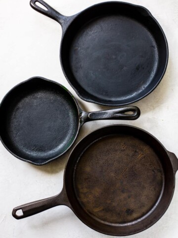 Three cast iron skillets of varying sizes are arranged on a white surface. Two have handles on one side, while the largest has two handles. Each skillet shows signs of use, revealing how to properly season a cast iron pan for lasting performance.