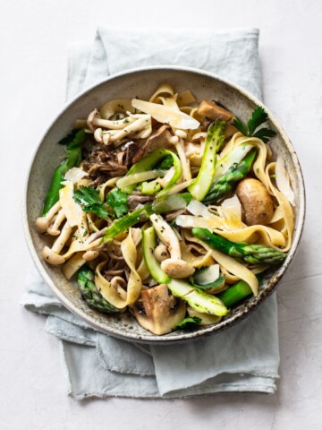 A bowl of asparagus mushroom pasta with tagliatelle, fresh parsley, and shaved parmesan, served on a light gray cloth napkin against a white background.