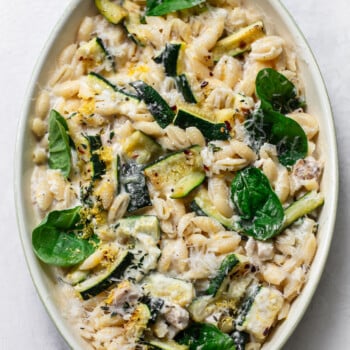 Oval baking dish filled with creamy chicken pasta, zucchini slices, fresh spinach leaves, and grated cheese, sitting on a light background.