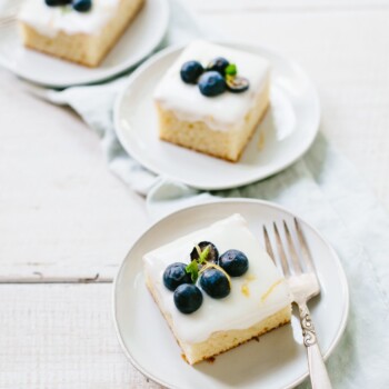 Three cake plates with slices of lemon sheet cake topped with creamy icing and fresh blueberries.