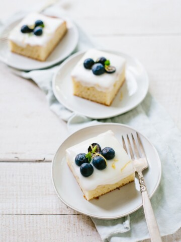 Three cake plates with slices of lemon sheet cake topped with creamy icing and fresh blueberries.