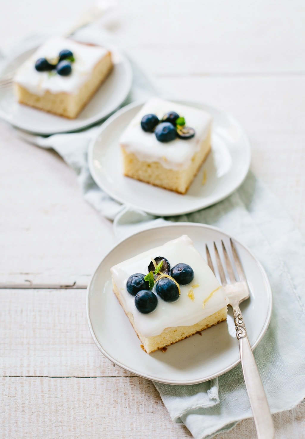 Three cake plates with slices of lemon sheet cake topped with creamy icing and fresh blueberries.