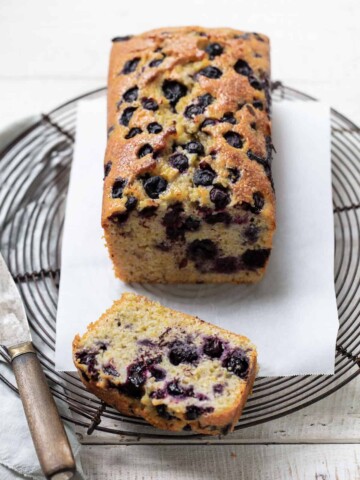 A blueberry loaf cake rests on a cooling rack lined with parchment paper. One slice is cut and laid in front, revealing blueberries inside. A knife sits nearby on the white wooden surface.