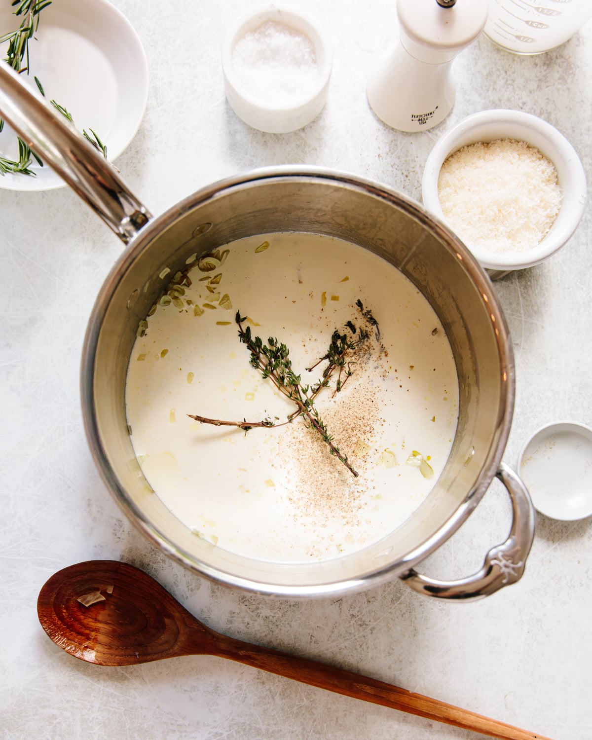 A saucepan filled with cream, garlic, thyme sprigs, and black pepper sits on a white surface, surrounded by bowls of salt, grated cheese, chopped potatoes, a wooden spoon, and a pepper grinder.