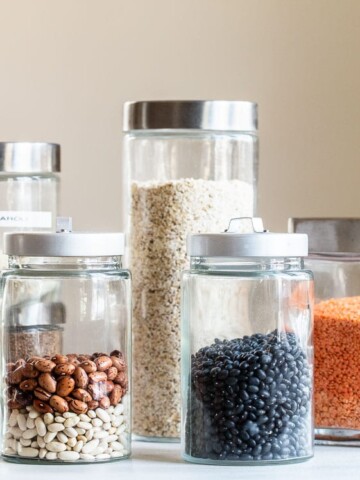 Six glass jars of various sizes, filled with pantry staples like beans, lentils, grains, and seeds, are arranged on a white surface against a neutral beige background. Each jar has a metal lid.