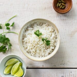A bowl of cooked coconut rice garnished with cilantro sits on a light wooden surface. Nearby are a small bowl of cardamom pods, lime wedges in a dish, and a sprig of cilantro.