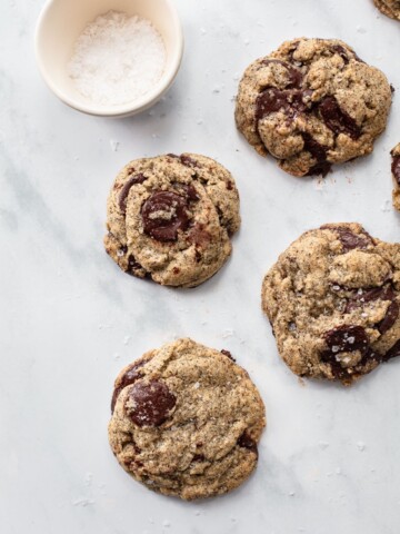 Five buckwheat chocolate chip cookies are scattered on a light marble surface, with a small white bowl of coarse sea salt in the upper left corner. Some cookies are sprinkled with salt.