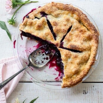 A homemade blueberry pie with a golden crust sits partially sliced in a glass dish on a white wooden table, surrounded by pink flowers and a pink napkin. A pie server rests inside the dish.