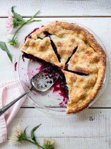 A homemade blueberry pie with a golden crust sits partially sliced in a glass dish on a white wooden table, surrounded by pink flowers and a pink napkin. A pie server rests inside the dish.