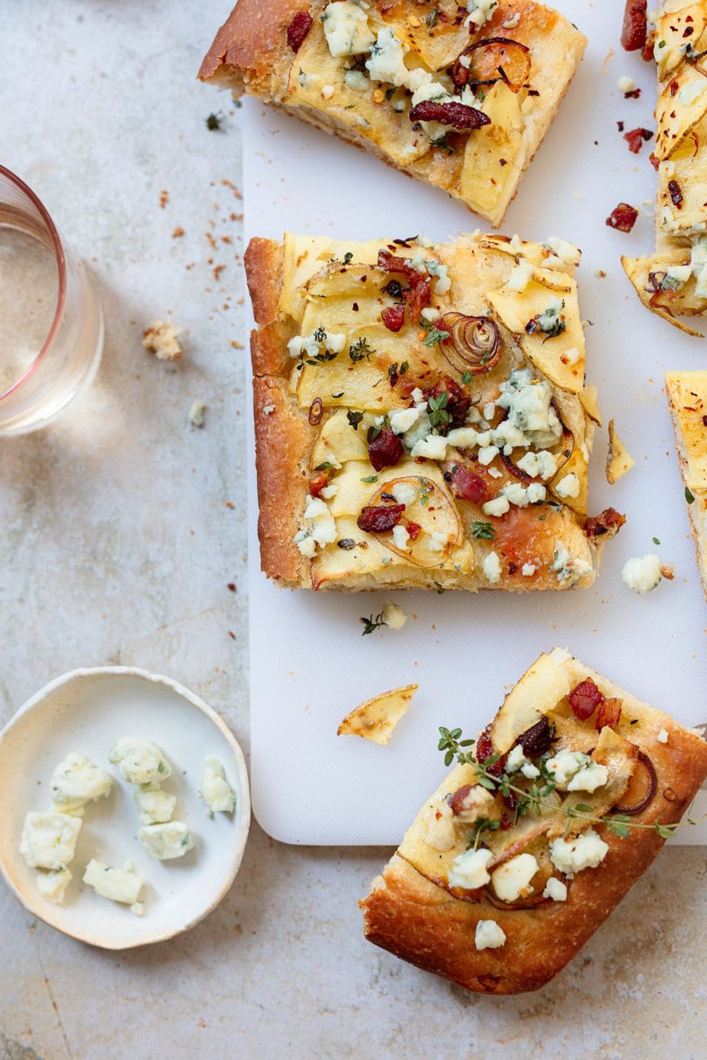 Rectangular slices of potato focaccia topped with cheese, thinly sliced potatoes, herbs, and small pieces of bacon are arranged on a white cutting board, with a small plate of crumbled cheese and a glass of water nearby.