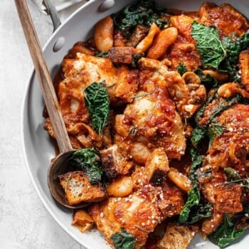 A skillet filled with chicken with white beans, tomato sauce, kale, and crispy bread cubes, garnished with grated cheese and fresh herbs. A wooden spoon rests on the side of the pan.