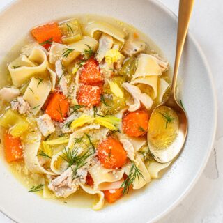 A bowl of chicken noodle soup with chunks of carrots, celery and chopped dill on top.