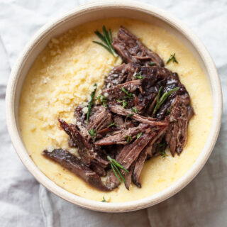 A bowl of creamy polenta topped with tender red wine short ribs, garnished with fresh rosemary and grated cheese, on a light fabric background.