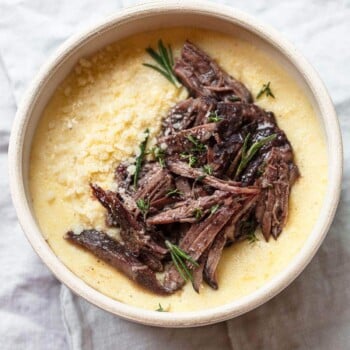 A bowl of creamy polenta topped with tender red wine short ribs, garnished with fresh rosemary and grated cheese, on a light fabric background.