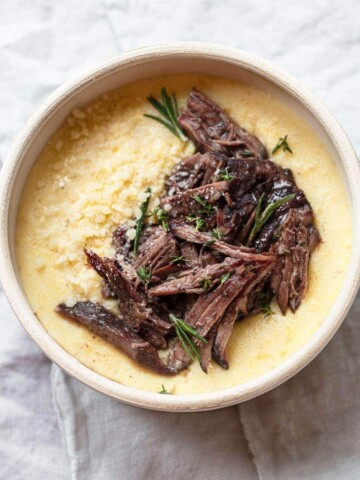 A bowl of creamy polenta topped with tender red wine short ribs, garnished with fresh rosemary and grated cheese, on a light fabric background.