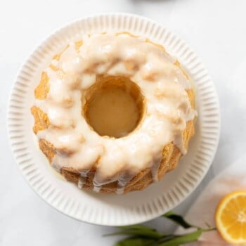 A glazed cake on a white plate with fresh Meyer lemon halves and leaves