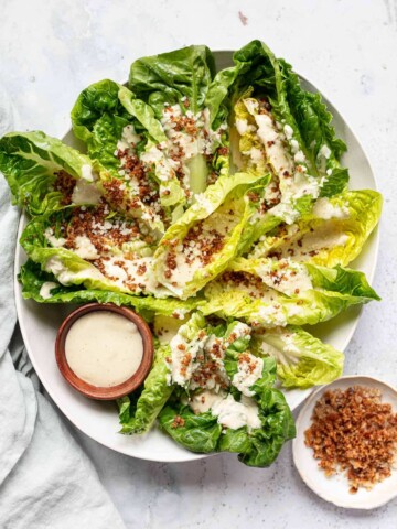 A plate of romaine lettuce leaves topped with creamy tahini caesar salad dressing, crumbled cheese, and crispy bits, served with a small bowl of extra dressing and a side dish of more crispy topping.
