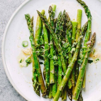 A white plate with roasted asparagus spears garnished with chopped green onions and a vinaigrette dressing, sitting on a light gray surface.