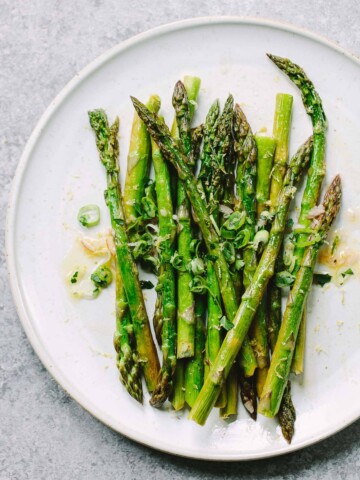 A white plate with roasted asparagus spears garnished with chopped green onions and a vinaigrette dressing, sitting on a light gray surface.
