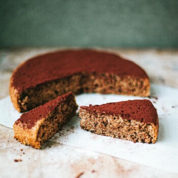 A round chocolate walnut cake dusted with cocoa powder sits on parchment paper. Two triangular slices have been cut and rest in front of the cake on a rustic wooden surface.