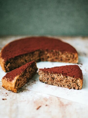 A round chocolate walnut cake dusted with cocoa powder sits on parchment paper. Two triangular slices have been cut and rest in front of the cake on a rustic wooden surface.