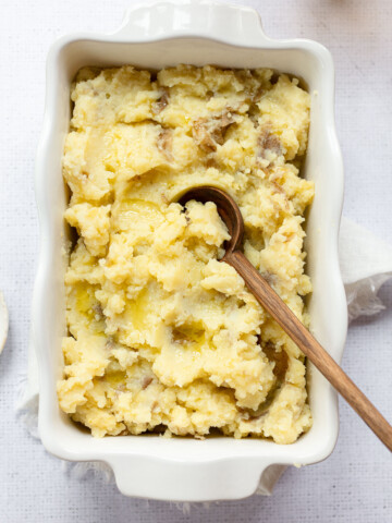 A white rectangular dish filled with olive oil mashed potatoes, topped with olive oil and a wooden spoon resting inside the mash. The potatoes appear rustic with some skin and chunks visible.