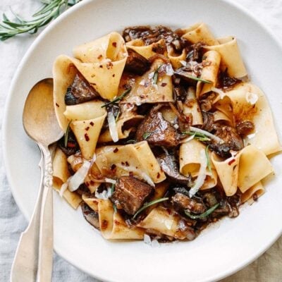 A bowl of mushroom pappardelle tossed with chunks of thick mushrooms in a dark savory sauce, topped with shaved cheese and fresh rosemary, with a silver spoon on the side on a light linen tablecloth.