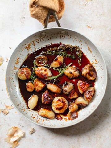 A pan with caramelized cipollini onions in a rich brown glaze, garnished with fresh rosemary and thyme, sits on a light textured surface with some garlic skins scattered nearby.
