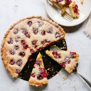 A frangipane tart topped with raspberries and a crumbly crust, dusted with powdered sugar. One slice is being lifted out, and two slices are served on a plate with two forks. Crumbs are scattered on the white surface.