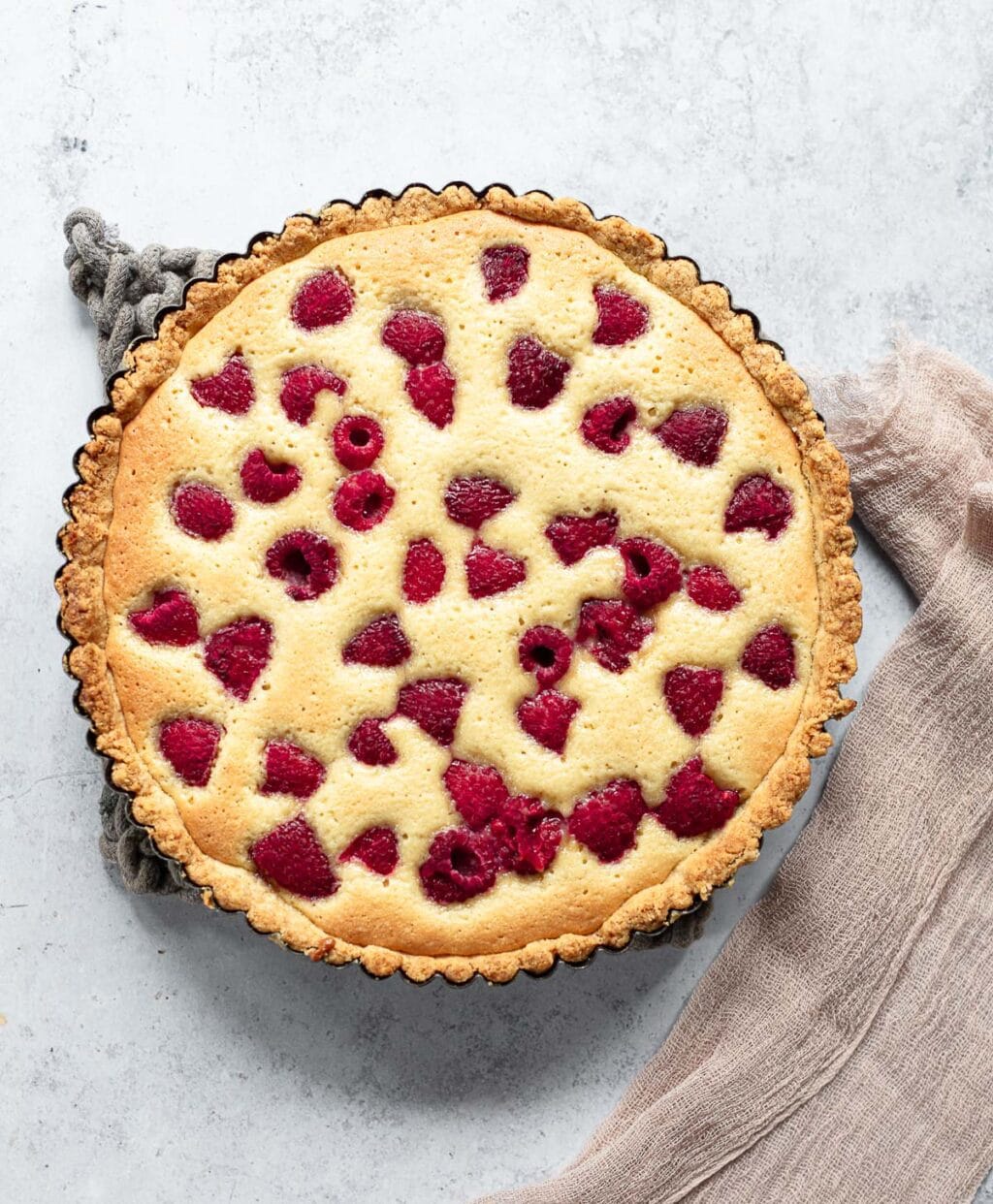 A baked frangipane tart with a golden crust, topped with scattered fresh raspberries, sits on a textured gray surface with a beige cloth napkin beside it.