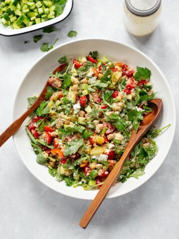 A large white bowl of colorful bulgur wheat salad with chopped tomatoes, cucumbers, chickpeas, cilantro, and greens—like a vibrant vegetable tabbouleh—is served with wooden salad tongs. A dish of diced cucumbers and a jar of dressing are nearby.