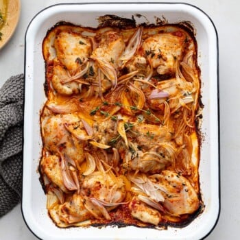 A white baking dish filled with baked boneless chicken thighs, sliced shallots, and fresh herbs in a golden, seasoned sauce. The dish is set on a light surface with part of a gray cloth visible to the side.