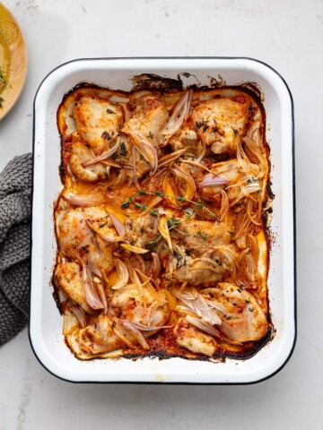 A white baking dish filled with baked boneless chicken thighs, sliced shallots, and fresh herbs in a golden, seasoned sauce. The dish is set on a light surface with part of a gray cloth visible to the side.
